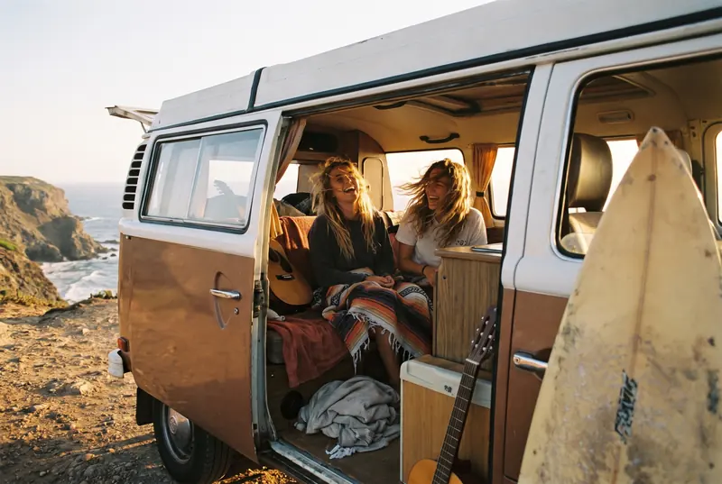 Friends sitting in an open camper van on the coast, a surfboard leaning beside it